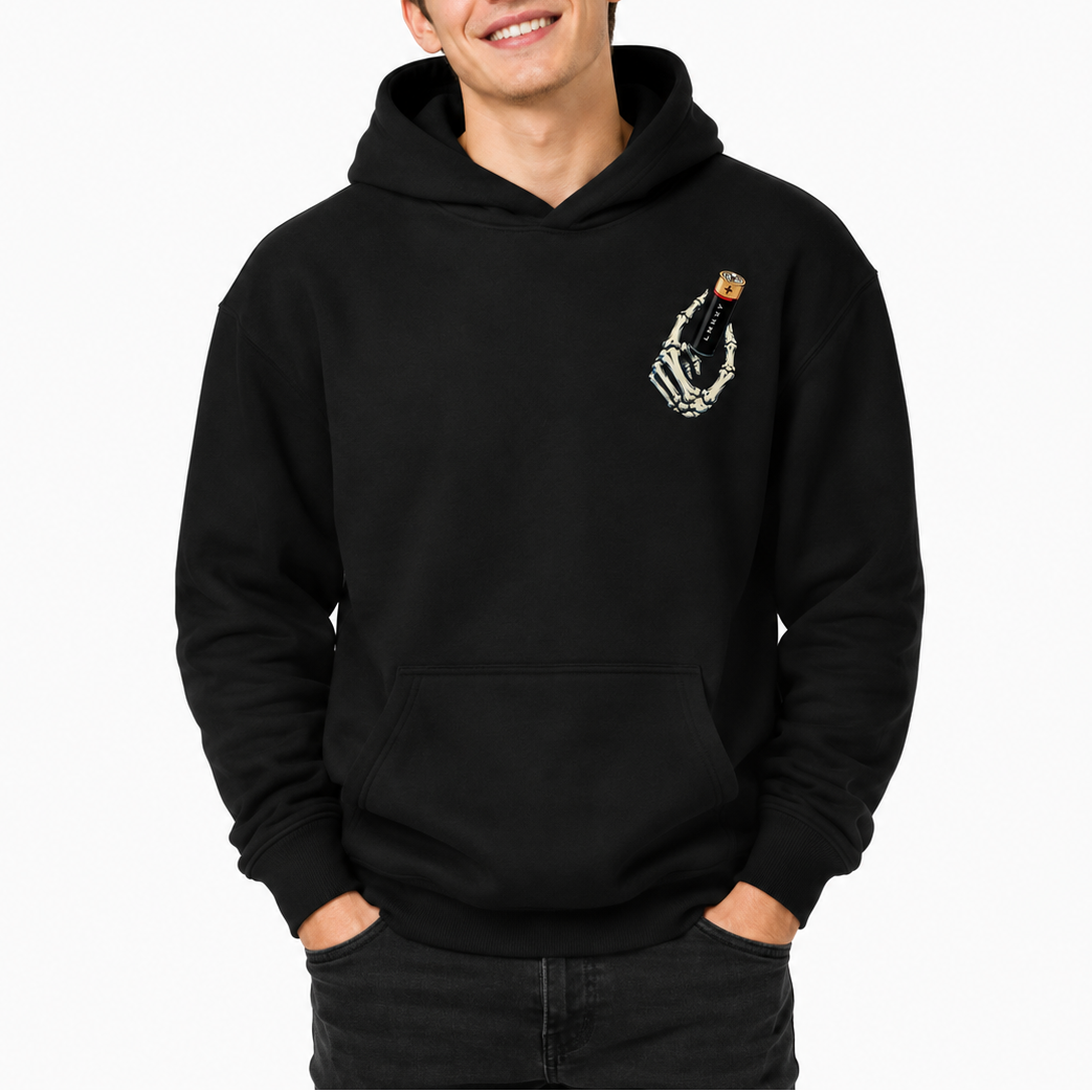 A smiling young man wearing a black pullover hoodie with a small white skeleton hand graphic holding a gold cup on the left chest, paired with dark jeans against a plain white background.